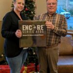 Two individuals smiling and holding an AED box labeled “Encore Series AEDs - Because We Believe in Second Chances,” standing in front of a Christmas tree with wrapped gifts at the base.