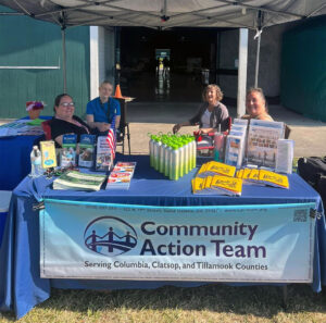 A group of four Seniors Program staff from Community Action Team (CAT) are seated at a booth at the Columbia County Fair. The booth is covered with informational brochures, water bottles, and other materials. A large banner in front displays the CAT logo and their service areas: Columbia, Clatsop, and Tillamook Counties.